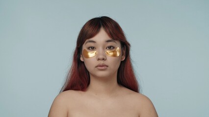 Close-up isolated photo of a young seminude woman with red dyed hair and nude makeup wearing two golden eyepatches under her eyes. - obrazy, fototapety, plakaty