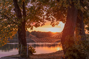 Riverbank with hammock for relaxing on a warm fall evening at sunset with backlit red sun, a place to relax in nature