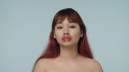 Close-up isolated photo of a young seminude woman with red dyed hair and nude makeup wearing a lip patch, looking at the camera, relaxed. - obrazy, fototapety, plakaty