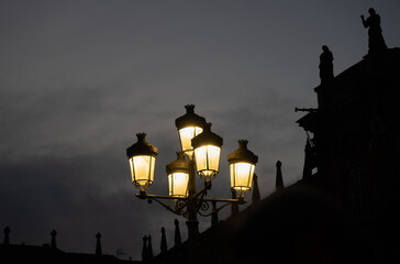 Wrought iron lamp with five lanterns dimly illuminating with yellow light at dusk in the main square of Salamanca with the gray and dramatic sky and the silhouette of the palace and two sculptures