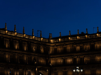 Facade of houses dimly lit with wooden windows and the warm light of the baroque facade of the main square of Salamanca with the dark blue sky at dusk.