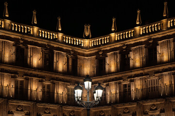 Wrought iron lamp with five lanterns dimly illuminating the main square of Salamanca with the sky completely dark and the palace dimly illuminating the wooden windows and baroque façade warm light. © sirbouman