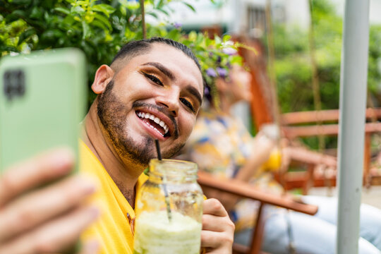portrait of a homosexual man with makeup taking a very happy picture with his cell phone and having a drink and behind him his short-haired friend with purple hair smiling 