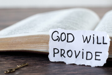 God will provide, handwritten quote on paper in front of open holy bible book. Close-up. Care,...