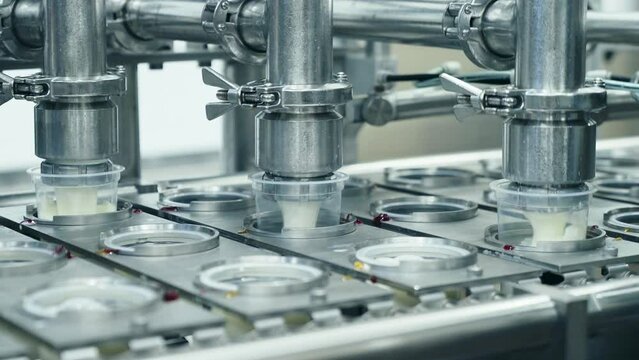 Automated machine is filling up the yogurt containers at the dairy facility. Industrial machine is used for filling the cups with the yogurt. Machine is pouring the yogurt to fill up the containers.