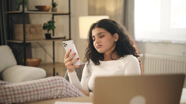 Boring exhausted young woman student or freelancer scrolling feed of social networks products in internet store on smartphone instead of work or education at home workplace Procrastination concept