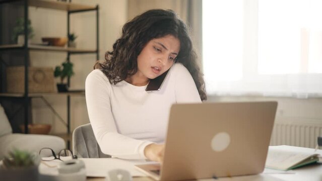 Nervous stressed young woman freelancer typing quickly on laptop computer hurry up scream and warning about deadline while having working call conversation by smartphone at light home workplace