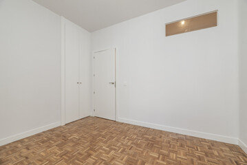 Empty room with a small new built-in wardrobe with white wooden sliding doors, access door of the same material, skylight on the white wall and reddish parquet floor