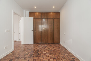 An empty room with a large built-in wardrobe with sapelly-colored wooden doors covering one wall and reddish parquet floors