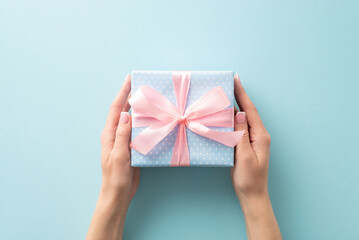Wrapped with love: woman's hands from first person top view hold a pastel blue gift box, featuring polka dots and a pink ribbon bow, on a pastel blue canvas, leaving room for your text or advert