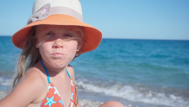Close-up of a girl in a hat turning to the camera and blowing an air kiss. A happy girl is sitting on the shore in a hat.