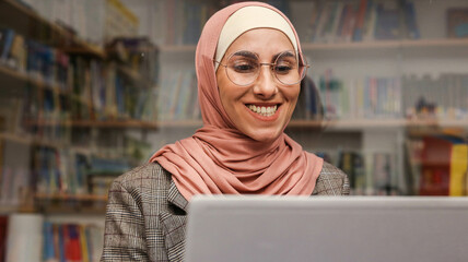 Close-up portrait beautiful Muslim woman in hijab with glasses sitting in library working at laptop...