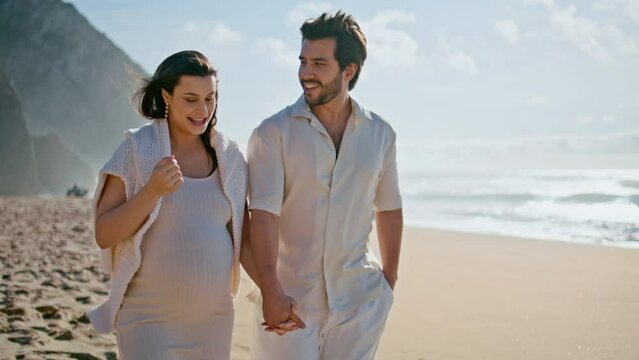 Pregnant woman walking husband on sunny ocean shore talking laughing together.