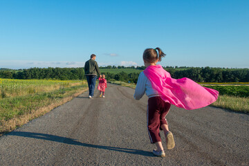 Сhild in a superhero cape runs along the road to his dad with child. - 653453226