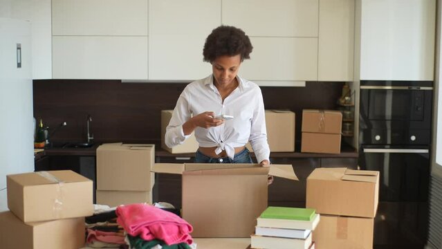 Front view of happy African-American woman leaving apartment, taking photo on smartphone of packing box for moving, ordering courier, delivery, shipping service, standing at cardboard container.
