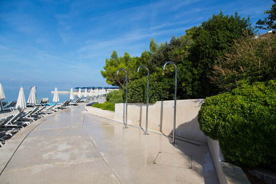 Garden design and landscaping: Showers, comfortable sun loungers and shady parasols at a a pine tree seamed promenade with elaborate nature stone paving slabs at a pebbles beach, Rovinj, Croatia