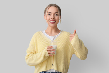 Young woman with glass of milk showing thumb-up on grey background