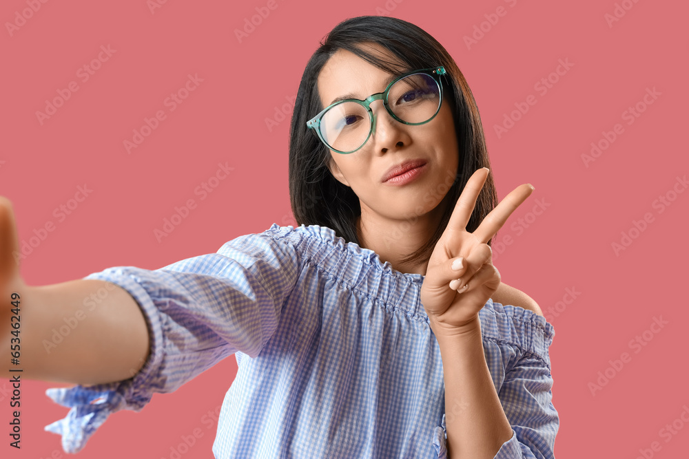 Wall mural Beautiful Asian woman in stylish eyeglasses taking selfie on pink background