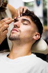 Obraz premium Vertical photo of a bearded young adult with his eyes closed is sitting in a beauty center cutting his eyebrows.The woman stylist uses tweezers to pull hairs from the eyebrows.Removal of eyebrow hairs