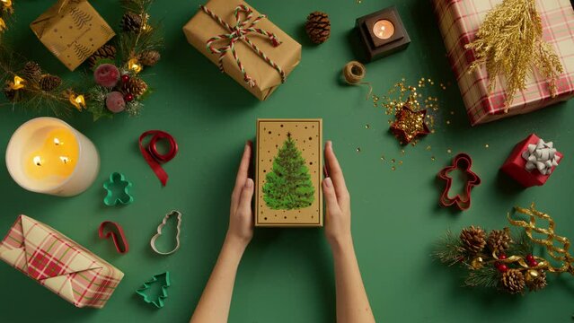Overhead female hands opening green and golden gift box with christmas tree drawing on green background. Presents wrapped in kraft paper for new year celebration. Holiday shopping background top view