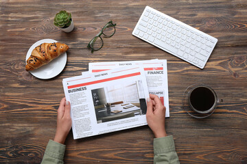 Woman reading newspaper at workplace with cup of coffee and croissant on wooden background