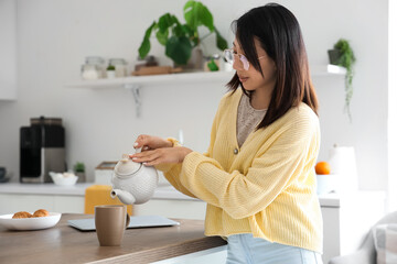 Beautiful young Asian woman pouring tea from teapot into cup at kitchen