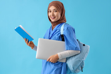 Female Muslim medical intern with laptop and book on blue background