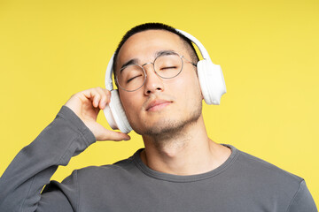 Portrait of pensive asian guy wearing stylish eyeglasses listening to music in wireless headphones