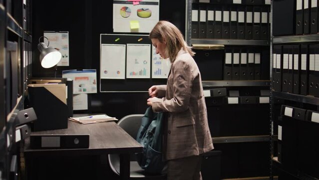 Caucasian detective inspector examines case files while seated at desk with laptop. Female private investigator carrying backpack enters police department office space and goes through crime records.
