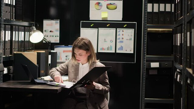 Private investigator enters office with case folder, examining evidence on desk with files and laptop. Caucasian woman with expertise in criminal investigation, reviews statements to identify suspect.