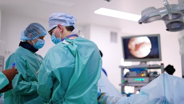 Rear view of surgeons in the operational room. Nurse passes the tool to a doctor. Screen works at the backdrop.