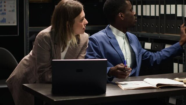 Multiethnic private detectives collaborate in incident room, conducting criminal investigation. African american and caucasian inspectors collecting clues and information using, laptop and case files.
