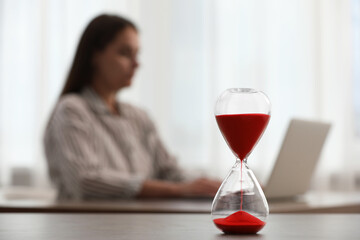Hourglass with red flowing sand on table. Woman using laptop indoors, selective focus