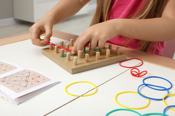 Fototapeta premium Motor skills development. Girl playing with geoboard and rubber bands at white table indoors, closeup