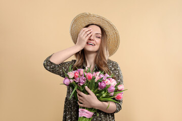 Happy young woman in straw hat holding bouquet of beautiful tulips on beige background