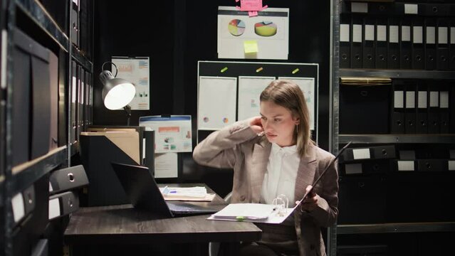 Detective walks inside office with case folder, scrutinising evidence on desk with papers and laptop. Woman with experience in criminal investigation examines statements to identify suspect. Zoom-in.