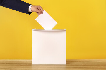 Man putting his vote into ballot box on yellow background, closeup