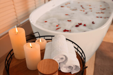 Burning candles, towels on table near bath tub with foam and rose petals in bathroom