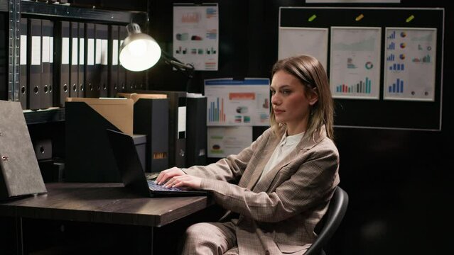 Caucasian private detective inspecting evidence, reviewing files, and using laptop in office. Side-view of female law officer checking for criminal files and records on minicomputer. Tripod shot.