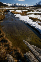 Arroyo de deshielo en invierno con troncos, agua limpia, nieve y tundra en primer plano y los Andes...