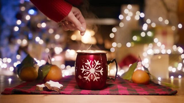 Female hand placing tea packet into cup with a hot water in red glossy mug with white snowflake on a wooden table with plaid napkin against blurred background of burning fireplace and Christmas tree