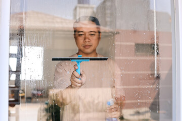 Asian cleaner man using detergent and squeegee handle while cleaning window
