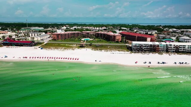 Destin FL drone shot flying sideways over the Gulf of Mexico with a view of the white sand, emerald green water, pompano joes restaurant, golf cart, parking lot, beach chairs, and umbrellas 2 of 2