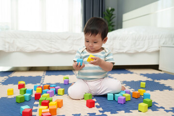 happy infant baby playing wooden block toy on jigsaw mat in bedroom