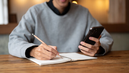 A young Asian man using his smartphone and taking notes on his book while sitting in a coffee shop.