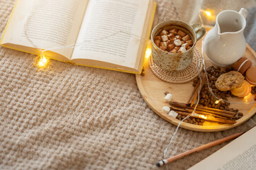 Top view of an opened book, a hot cocoa mug, and accessories are on a bed in a cosy bedroom.