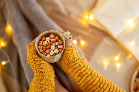 A woman in a cosy sweater and winter socks sitting on a bed with a cup of hot cocoa in her hands.