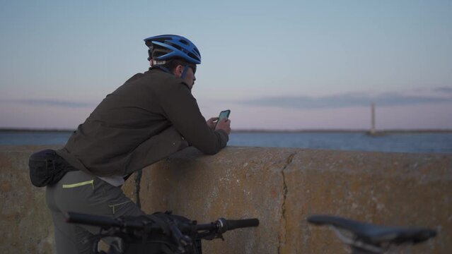 Theme is cycling in seaside. Male cyclist stopped to enjoy the sea view and see the road on the map on his smartphone. A male with a bicycle on the embankment is resting and using a smart phone.