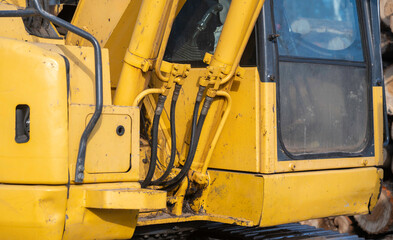 Closeup detail of a working, used, dirty yellow excavator, centered on hydraulics hoses and pipes next to the window of the operator cab.