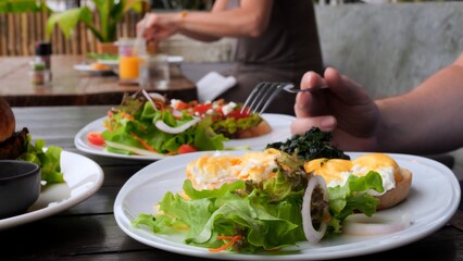 Man eating full meal as poached egg, salad and avocado toast in cafe. Close-up of man with fork pierces poached egg and eat tasty yolk. Order poached egg to eat healthy full meal. Full meal concept.